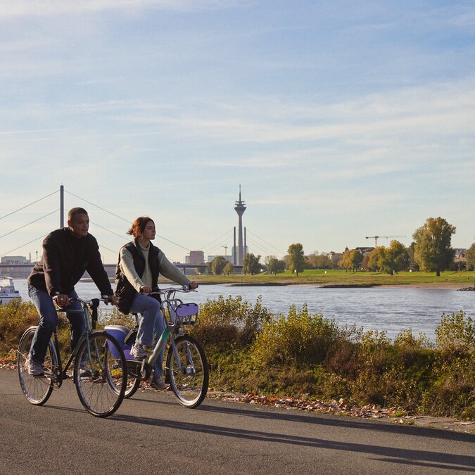 Two people cycle along the Rhine river with a view of the Rhine Tower. | © Düsseldorf Tourismus – React-EU