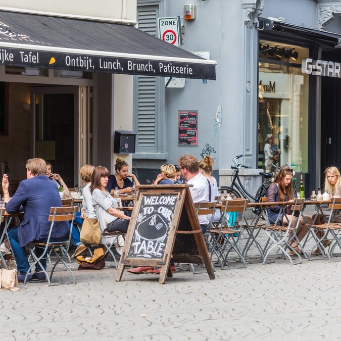 Busy terrace of a café in Ghent with guests sitting at wooden tables on a cobblestone street. | © Piet de Kersgieter – Terrace in Ghent