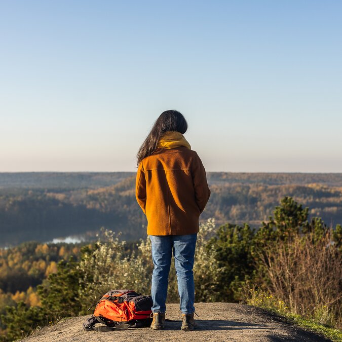 A person stands on a rocky outcrop overlooking a vast landscape of forests and lakes; a backpack lies on the ground next to the person. | © MEDIALIFE.BE