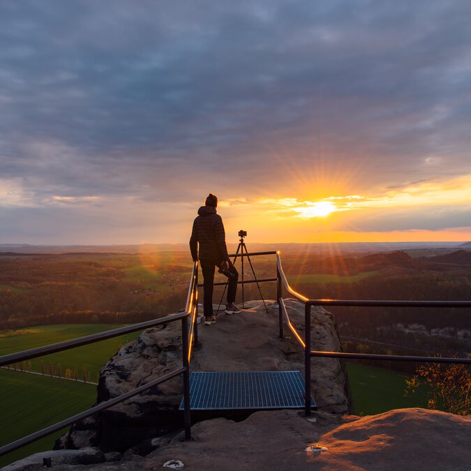 Person auf Aussichtspunkt betrachtet Sonnenuntergang über einer weiten Landschaft | © TMGS/CzechVibes