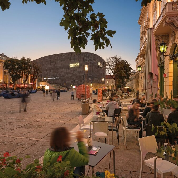 Vue sur une place au crépuscule depuis un café, avec en arrière-plan le mumok et la Halle du Museumsquartier à Vienne.  | © Wien Tourismus | Christian Stemper