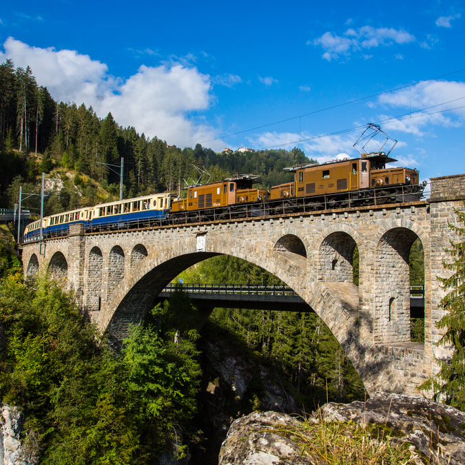 Le Glacier Pullmann Express, avec une locomotive brune et des wagons blancs et bleus, traverse un pont au milieu de la forêt, avec les montagnes et un ciel bleu derrière lui.