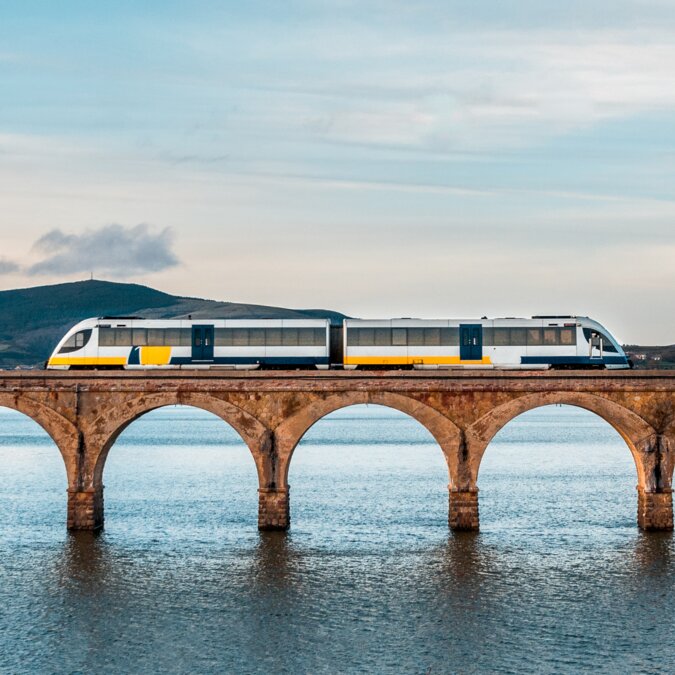 Rail journey through Spain with a modern train on a historic stone bridge over water, wide landscapes, and a calm atmosphere under a bright sky. | © shutterstock 1378378817